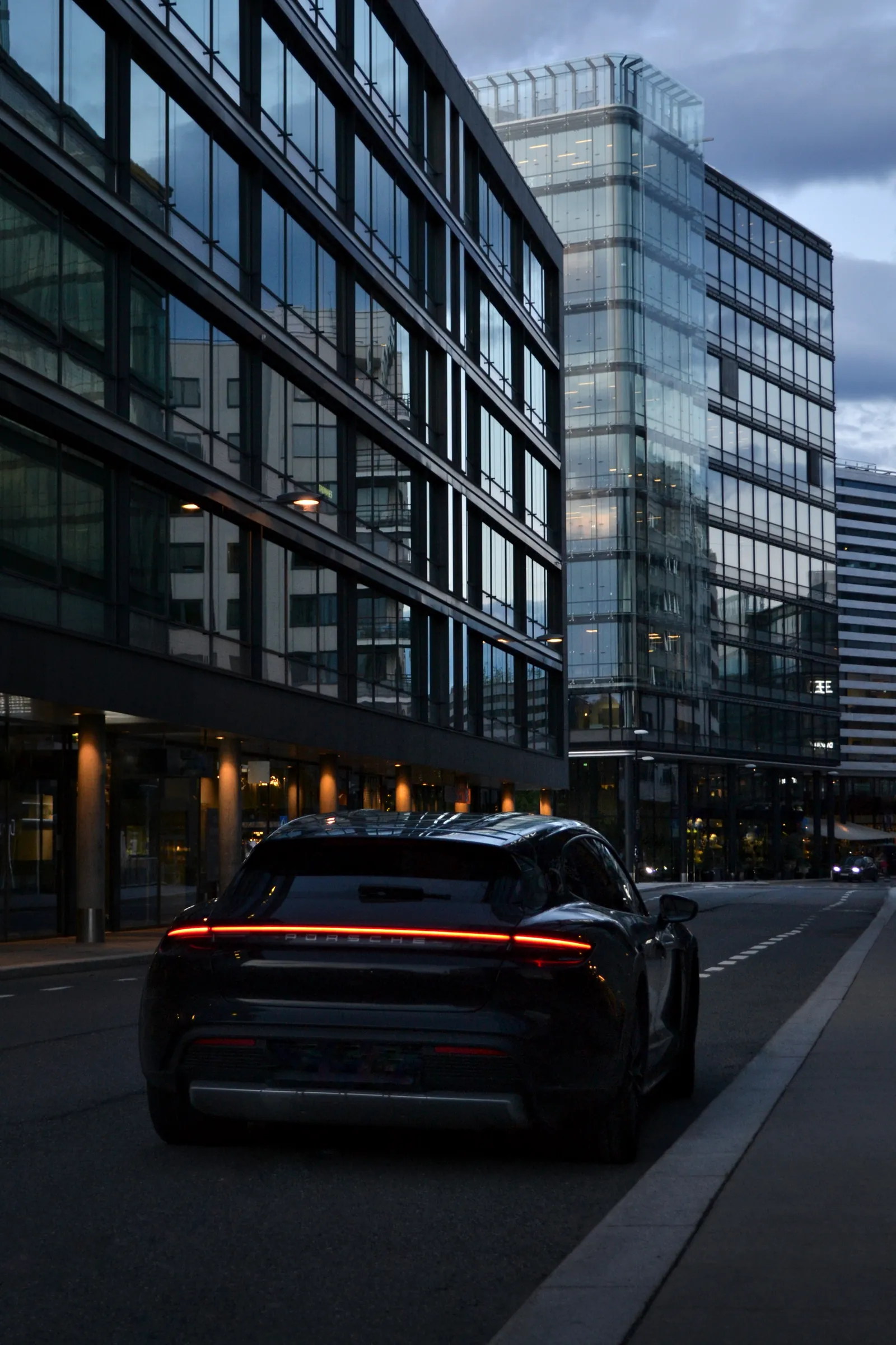 Dark Porsche with a light-bar tail lamp driving through a city street at dusk